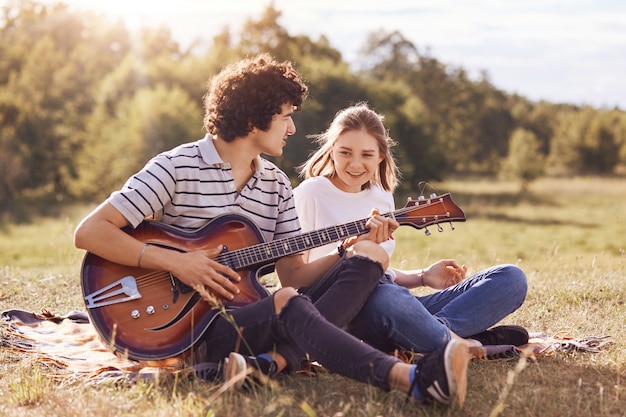 Photo d'amis heureux qui se divertissent, chantent des chansons et jouent de la guitare, ont des expressions joyeuses, s'assoient sur le sol, vêtus de vêtements décontractés, profitent d'une journée ensoleillée d'été sur la nature Concept de style de vie