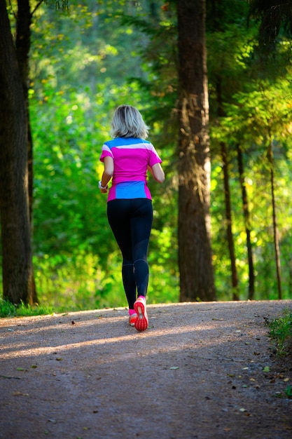 Photo de l'arrière d'une sportive blonde qui traverse un parc parmi les arbres le matin d'été.