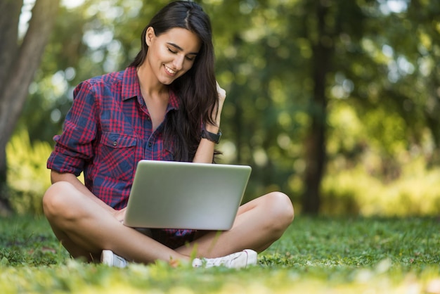 Photo attrayante d'une jeune femme de race blanche brune avec un sourire pigiste habillée décontractée assise sur la pelouse devant un ordinateur portable travaillant sur un ordinateur portable faisant du shopping lisant des articles étudiant