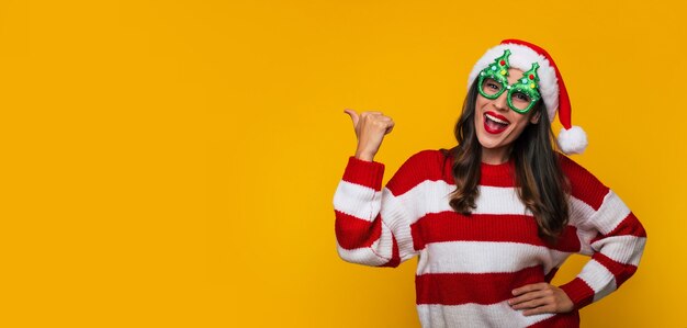 Photo de bannière d'une belle femme moderne excitée dans des lunettes de Noël amusantes et un bonnet de Noel s'amusant en posant contre un mur jaune