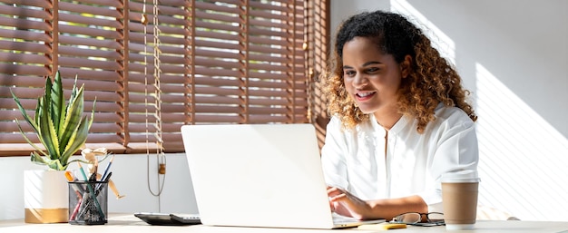 Photo de bannière d'une jeune femme d'affaires afro-américaine heureuse travaillant à domicile sur un ordinateur portable dans le salon