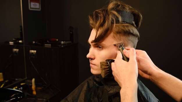 Photo d'un beau coiffeur donnant une coupe de cheveux à son client à l'aide d'une tondeuse Service de coiffeur dans un salon de coiffure moderne dans un éclair sombre avec une lumière chaude vue latérale