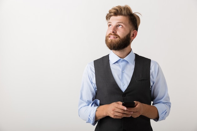 Photo d'un beau jeune homme barbu debout isolé sur fond de mur blanc à l'aide de téléphone mobile.