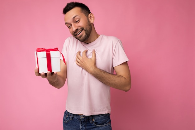 Photo d'un beau jeune homme brun surpris souriant heureux avec une barbe isolée sur un mur de fond rose portant un t-shirt rose tenant une boîte-cadeau blanche avec un ruban rouge et regardant le présent