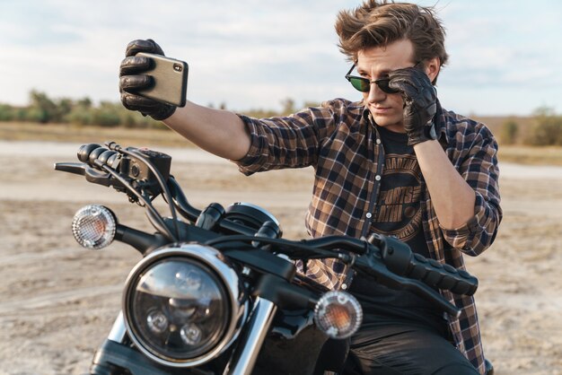 Photo d'un beau jeune homme motard à vélo à l'extérieur sur le terrain du désert prendre un selfie par téléphone portable.