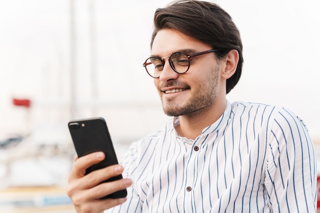 Photo d'un beau jeune homme portant des lunettes tapant sur son téléphone portable et souriant en marchant sur la jetée