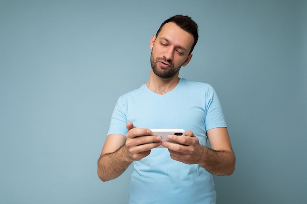 Photo d'un beau jeune homme sérieux avec une barbe portant un t-shirt bleu de tous les jours isolé sur fond bleu tenant et utilisant la communication par téléphone mobile en ligne sur Internet en regardant l'affichage gadjet