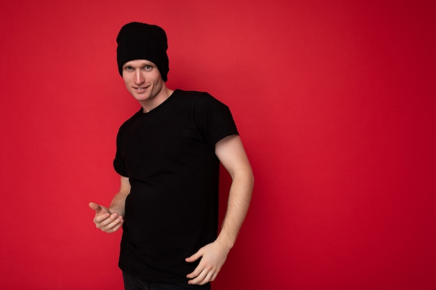 Photo d'un beau jeune homme souriant et heureux debout isolé sur un mur de fond rouge portant un t-shirt noir pour une maquette et un chapeau noir et regardant la caméra. Espace libre, espace de copie