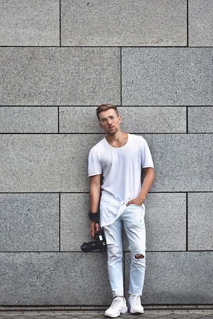 Photo D'un Beau Mec Caucasien Sur Un Mur Texturé Gris En T-shirt Blanc Et Jean Blanc Avec Caméra Accrochée à La Sangle Sur Son Bras.