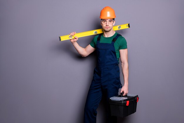 Photo de beau mec masculin ingénieur qualifié tenir mesure boîte à outils rack prêt à commencer le travail porter t-shirt salopette de sécurité casque de protection lunettes mur gris isolé