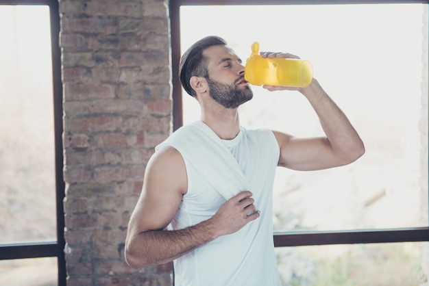 Photo de beau mec sportif fini la formation du matin à boire de l'eau douce tenant une serviette les yeux fermés sportswear débardeur formation maison windows à l'intérieur