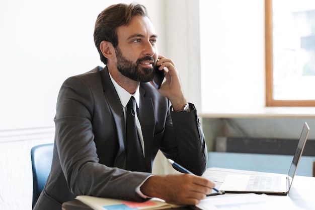 Photo d'un bel homme d'affaires barbu optimiste et heureux assis au bureau à l'aide d'un ordinateur portable travaillant à l'intérieur en parlant par téléphone portable.