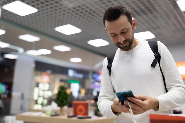 La photo d'un bel homme brune positif adulte avec du chaume dans un sweat-shirt blanc choisit un mobile