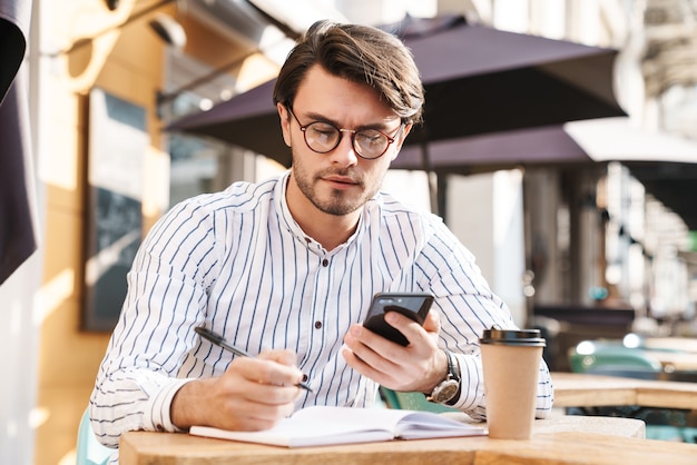 Photo d'un bel homme concentré portant des lunettes, tapant sur son téléphone portable et prenant des notes tout en travaillant dans un café en plein air