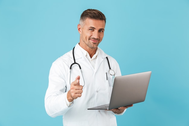 Photo de bel homme portant une blouse médicale blanche et un stéthoscope tenant un ordinateur portable, debout isolé sur un mur bleu