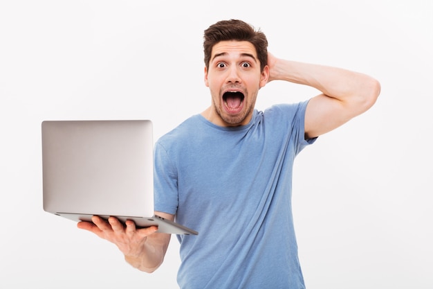 Photo de bel homme en t-shirt décontracté tenant un ordinateur portable argenté et regardant la caméra avec excitation ou choc, isolé sur mur blanc