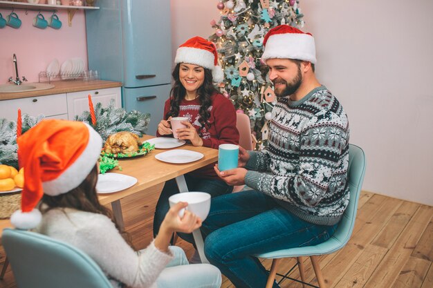 Photo d'une belle famille assise au tabel et se regarde. Ils tiennent des tasses dans les mains. Les gens se sourient. Il y a de la dinde et des mandarines sur la table.