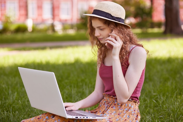 Photo de belle femme aux cheveux foxy assis sur l'herbe verte avec un ordinateur portable et un téléphone dans les mains, parler au téléphone