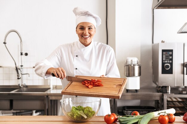 Photo de belle femme chef vêtu d'un uniforme blanc faisant de la salade avec des légumes frais, dans la cuisine au restaurant