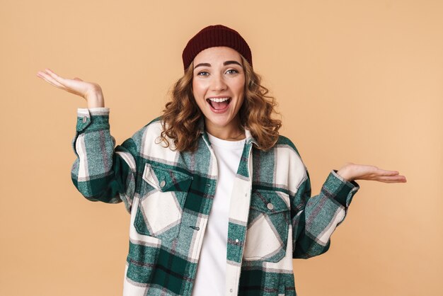 Photo d'une belle femme excitée en bonnet tricoté souriant et tenant un espace isolé sur beige