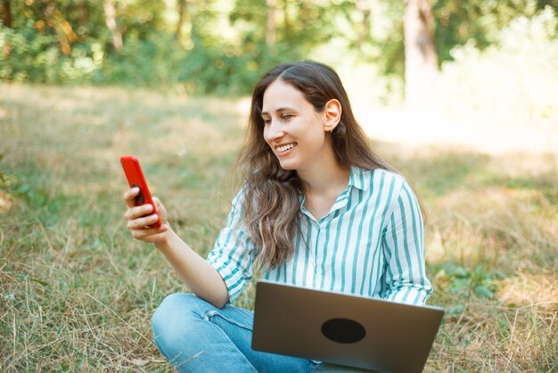 Photo de belle femme heureuse à l'aide de son téléphone dans le parc alors qu'il était assis sur l'herbe.