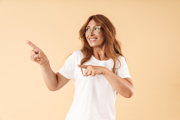 Photo d'une belle femme mature et heureuse souriante et souriante posant isolée sur un mur beige pointant vers le fond.