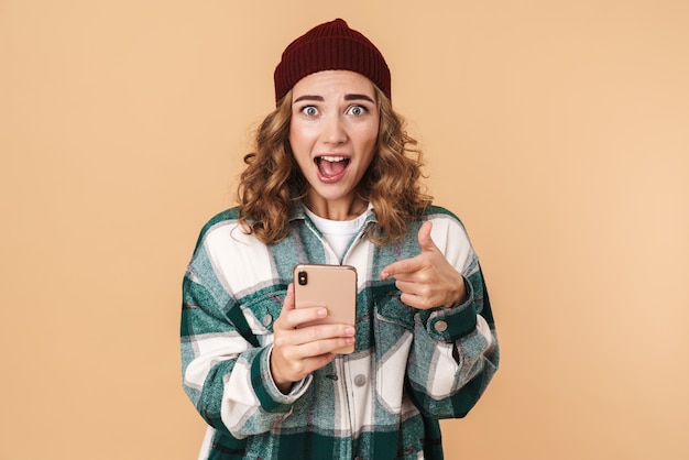 Photo d'une belle femme surprise en bonnet tricoté tenant et pointant sur un téléphone portable isolé sur beige