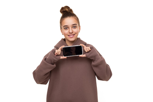 Photo d'une belle fille mignonne aux cheveux noirs et un chignon dans un sweat à capuche marron avec une capuche tenant un téléphone portable avec un écran noir vierge isolé sur fond blanc avec un espace libre pour le texte