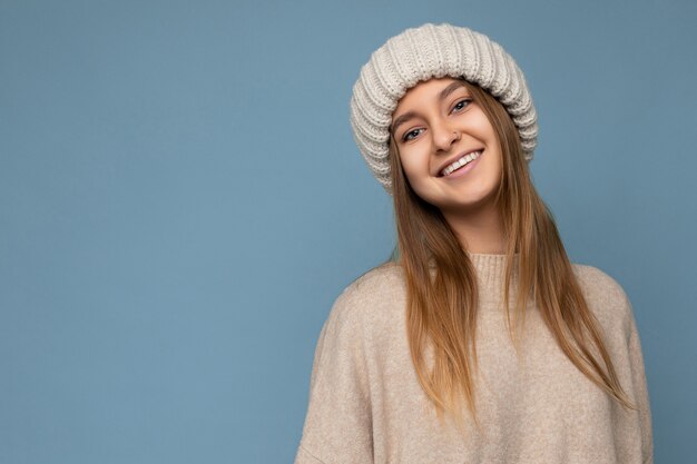Photo d'une belle jeune femme blonde foncée souriante et heureuse isolée sur un mur de fond bleu portant un pull chaud beige et un chapeau beige tricoté regardant la caméra. Espace libre, espace de copie
