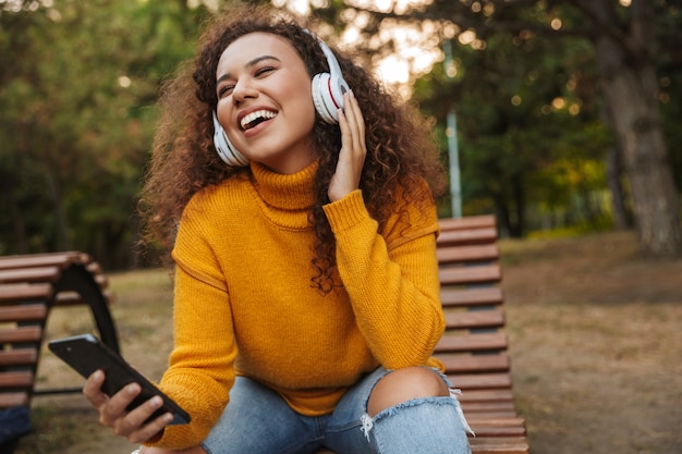 Photo d'une belle jeune femme bouclée optimiste joyeuse s'asseoir sur un banc dans un parc à l'extérieur en écoutant de la musique avec des écouteurs à l'aide d'un téléphone portable.