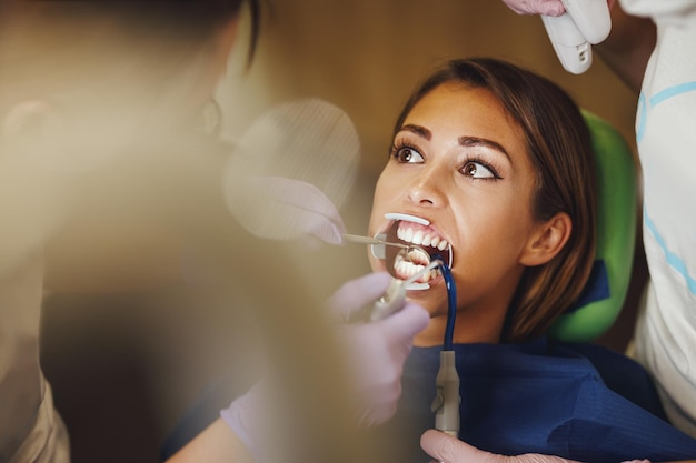 Photo d'une belle jeune femme chez le dentiste. Elle est assise dans le fauteuil du dentiste et le dentiste se prépare à mettre des accolades sur ses dents en mettant des serrures linguales auto-alignantes esthétiques.