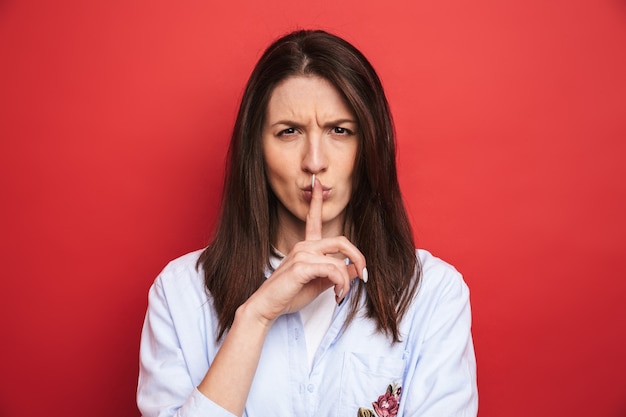 Photo d'une belle jeune femme en colère posant isolée sur un mur rouge montrant un geste de silence.