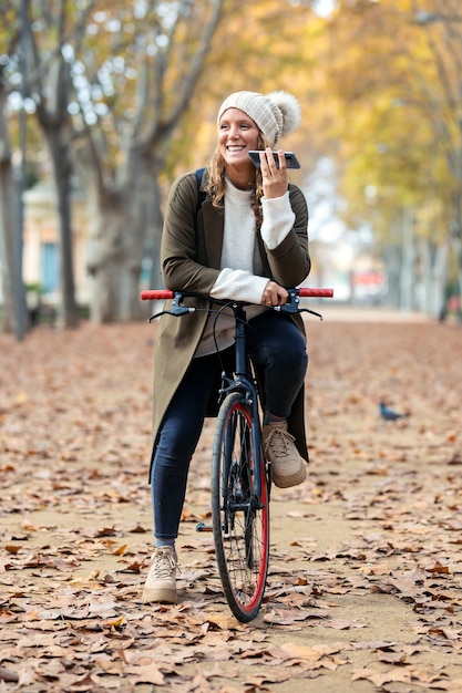 Photo photo d'une belle jeune femme envoyant un message audio vocal avec son téléphone intelligent tout en faisant du vélo dans le parc en automne.