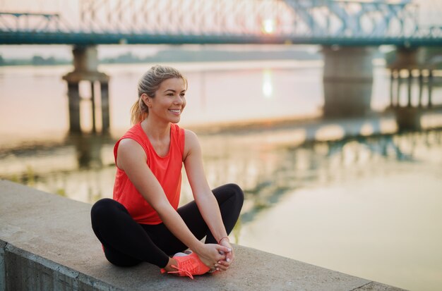 Photo de la bonne jeune femme en forme étirant ses muscles avant un entraînement intensif. Assis sur la bande de la rivière.