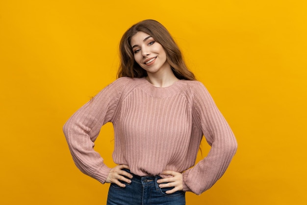 Cette photo capture la beauté naturelle d'une jeune femme aux cheveux longs Sur un fond jaune joyeux, son chandail confortable et sa posture détendue créent une ambiance de confort et de facilité