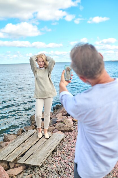 Photo chanceuse. Homme debout dos à la caméra avec un smartphone photographiant une femme posant joyeuse avec les mains levées près de la mer