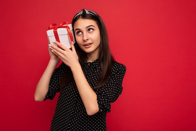 Photo d'une charmante fille brune réfléchie et heureuse isolée sur un mur de fond rouge portant un chemisier noir tenant une boîte-cadeau blanche avec un ruban rouge et levant les yeux. Espace libre