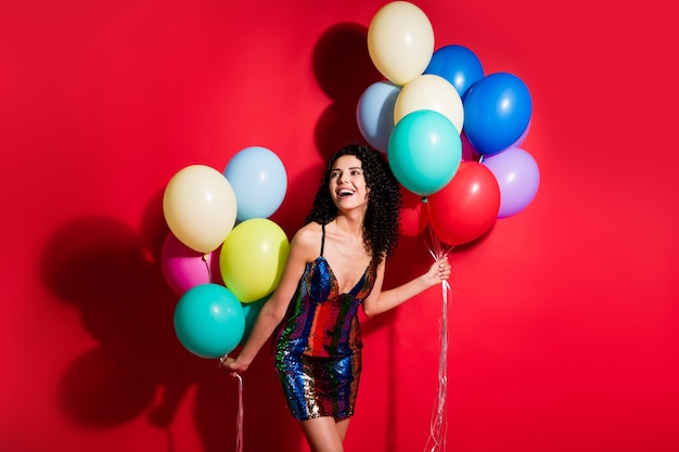 La photo d'une charmante fille tient de nombreux ballons à l'air de l'espace vide porte une robe courte brillante isolée sur fond de couleur rouge vif