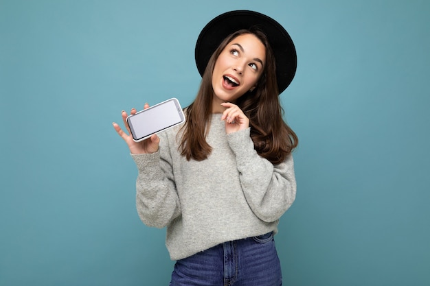 Photo d'une charmante jeune femme heureuse réfléchie portant un chapeau noir et un pull gris tenant un téléphone en levant isolé sur fond. Maquette, découpe, espace de copie