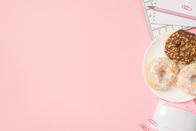 Photo ci-dessus d'un bloc-notes clavier souris d'ordinateur avec stylo trombones et assiette de beignets glacés isolés sur fond rose avec fond