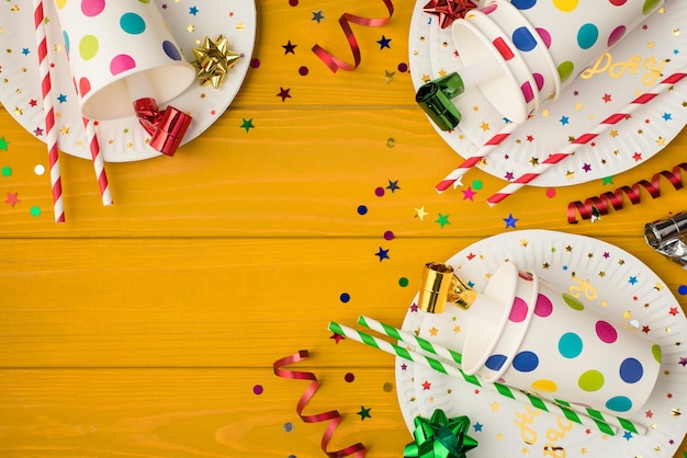 Photo ci-dessus de tasses à boire avec des tubules de fête sifflent un arc de confettis multicolores et des assiettes isolées sur le fond en bois jaune avec un espace vide
