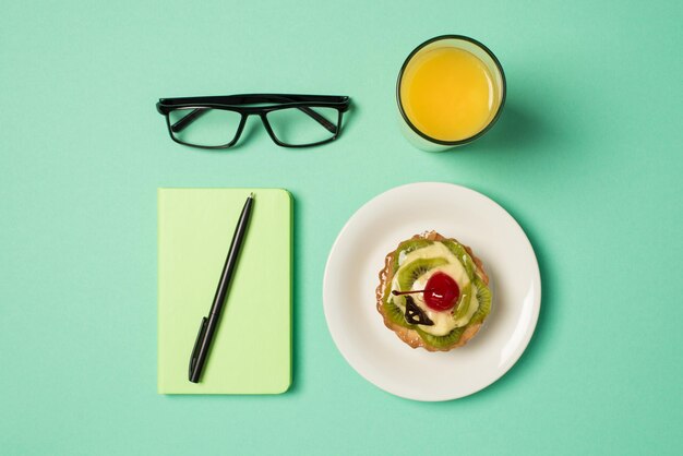 Photo ci-dessus d'un verre de jus de cahier vert avec des lunettes à stylo et une assiette avec un gâteau aux fruits isolé sur le fond bleu sarcelle