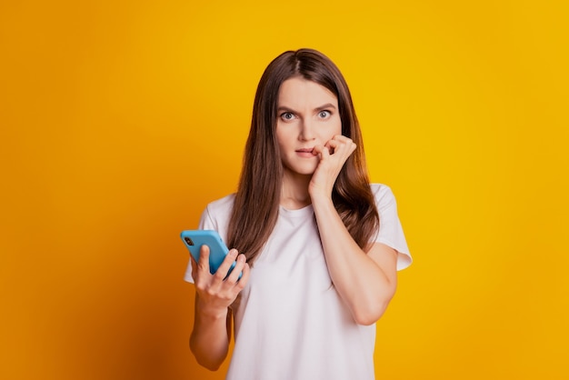 Photo de clous de morsure de dame inquiète tenir le téléphone porter un t-shirt blanc posant sur fond jaune