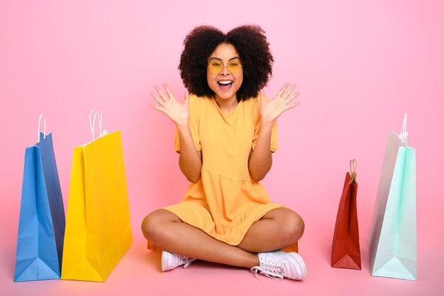 Photo complète d'une femme afro-américaine assez heureuse et bouclée en robe d'été orange assise entre des sacs à courses tient une carte bancaire de crédit regarde la caméra sourire isolé fond rose pastel