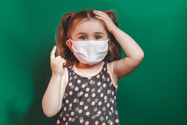 Photo conceptuelle de petite fille dans un masque médical montrant le doigt pointé sur fond vert bouchent 2020