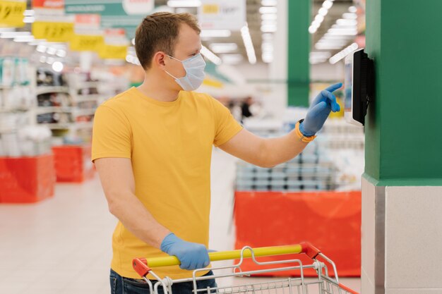 Photo d'un consommateur masculin qui utilise un écran tactile dans un magasin, vérifie le prix, pose avec son panier, porte un masque et des gants jetables