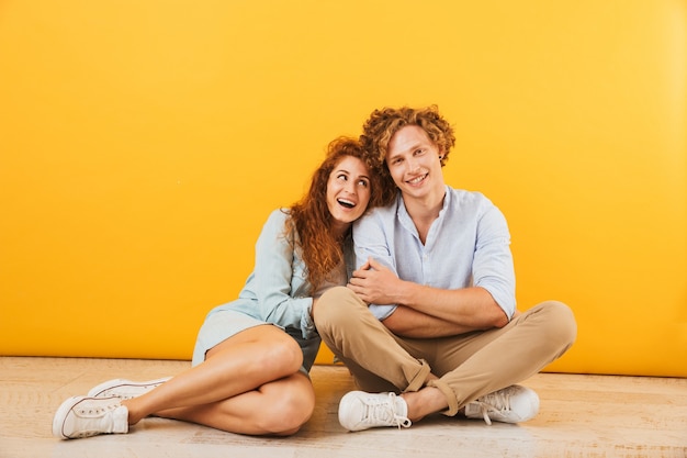 Photo de couple heureux optimiste homme et femme souriant et serrant assis sur le sol ensemble, isolé sur fond jaune