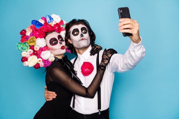 Photo de couple de zombies drôle mignon homme dame fille embrasser tenir téléphone faire selfie traduire festival porter robe noire costume de mort roses bandeau bretelles isolé fond de couleur bleu