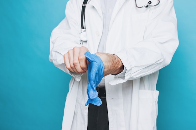 Photo de culture de l'homme médecin en blouse blanche met des gants médicaux en caoutchouc isolé sur fond bleu