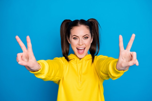 Photo d'une dame assez drôle, la coiffure à deux queues lève les deux bras montrant le symbole du signe v bonne humeur joyeuse dire salut les amis portent un pull à capuche jaune décontracté isolé sur fond de couleur bleu vif